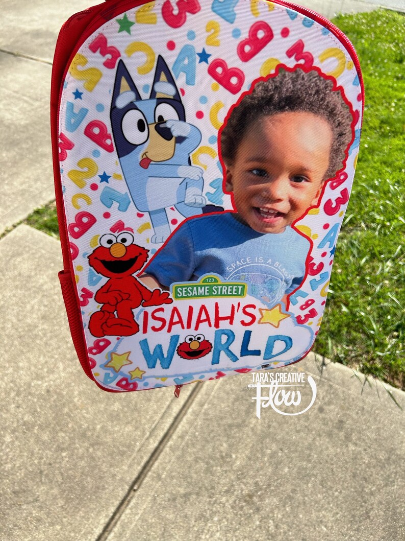 May include: A red backpack featuring a custom print with a smiling child, the cartoon character Bluey, and Elmo from Sesame Street. The background is white with colorful letters and numbers. The text reads Isaiah's World and Sesame Street.