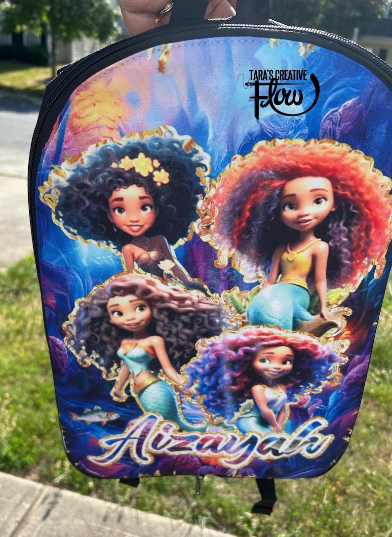 May include: A backpack featuring a vibrant underwater scene with four animated mermaid characters with curly hair. The design includes the text "Tara's Creative Flow" at the top and the name "Aizayah" in stylized blue and gold lettering at the bottom.