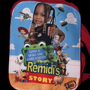 May include: A rectangular red lunch bag featuring a custom graphic print of a child surrounded by animated toy characters. The text reads: REMIDI IS... SMART, KIND, FUNNY & PRETTY, REMIDI'S STORY, and TARA'S CREATIVE FLOW. The background is blue.