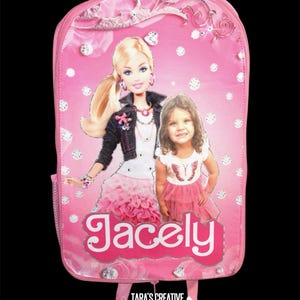 May include: A pink backpack featuring a graphic print of a blonde doll in a dark jacket and a young child in a white and pink outfit. The name Jacely is printed in white letters on a pink background. The design includes silver diamond-like accents.
