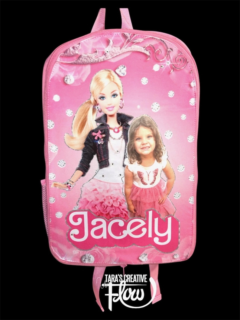 May include: A pink backpack featuring a graphic print of a blonde doll in a dark jacket and a young child in a white and pink outfit. The name Jacely is printed in white letters on a pink background. The design includes silver diamond-like accents.