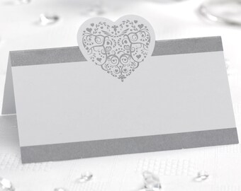 Puede incluir: Tarjeta de lugar blanca con un borde plateado y un diseño en forma de corazón plateado. El corazón tiene un patrón decorativo.