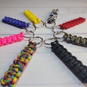 May include: A collection of eight paracord keychains arranged in a circle, featuring various colours including bright yellow, solid black, vibrant pink, red, purple and blue, camouflage, black and white, and a multi-coloured rainbow pattern.