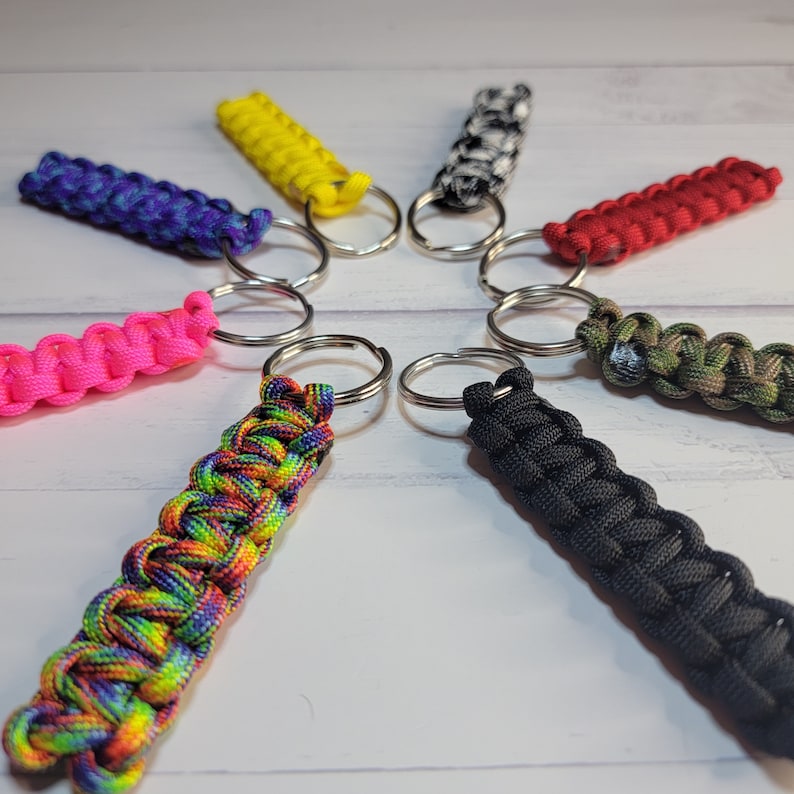 May include: A collection of eight paracord keychains arranged in a circle, featuring various colours including bright yellow, solid black, vibrant pink, red, purple and blue, camouflage, black and white, and a multi-coloured rainbow pattern.