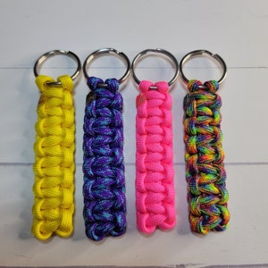 May include: Four keychains made of woven paracord, each attached to a silver metal split ring. The keychains feature different colours: one solid bright yellow, one purple and blue variegated, one solid hot pink, and one multi-coloured rainbow pattern.