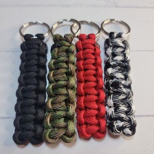 May include: Four paracord keychains featuring a cobra knot weave. The keychains are solid black, camouflage green and brown, solid red, and a black and white checkered pattern. Each keychain is attached to a circular silver metal split ring.