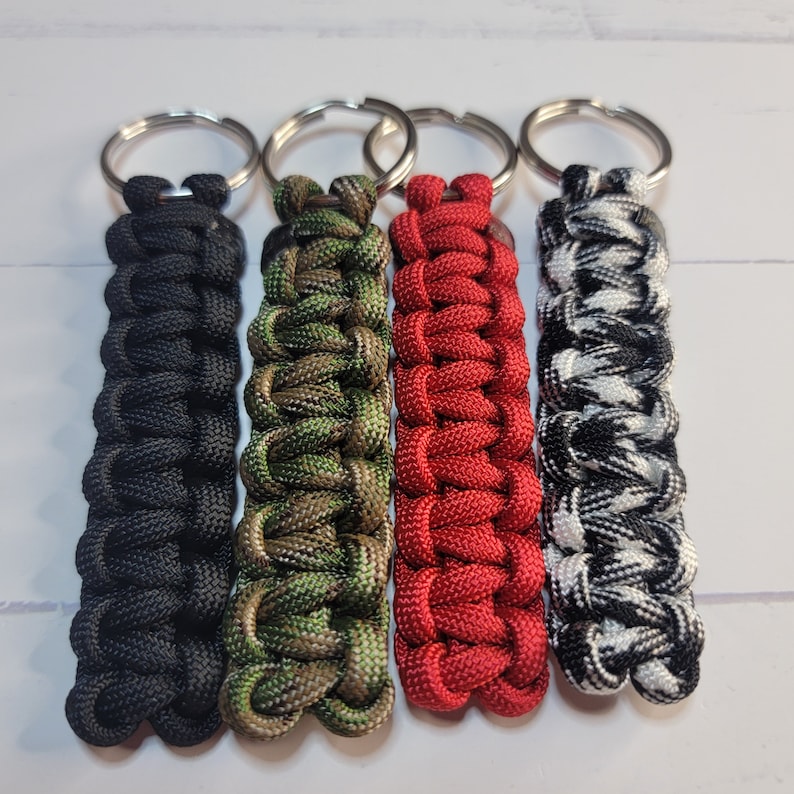 May include: Four paracord keychains featuring a cobra knot weave. The keychains are solid black, camouflage green and brown, solid red, and a black and white checkered pattern. Each keychain is attached to a circular silver metal split ring.