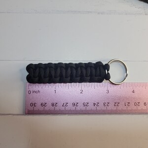 May include: A black paracord keyring featuring a woven cobra stitch pattern attached to a silver metal split ring. The keyring is positioned above a ruler, showing a total length of approximately 8.25 cm from end to end.