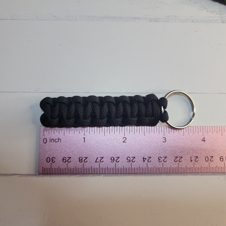 May include: A black paracord keyring featuring a woven cobra stitch pattern attached to a silver metal split ring. The keyring is positioned above a ruler, showing a total length of approximately 8.25 cm from end to end.