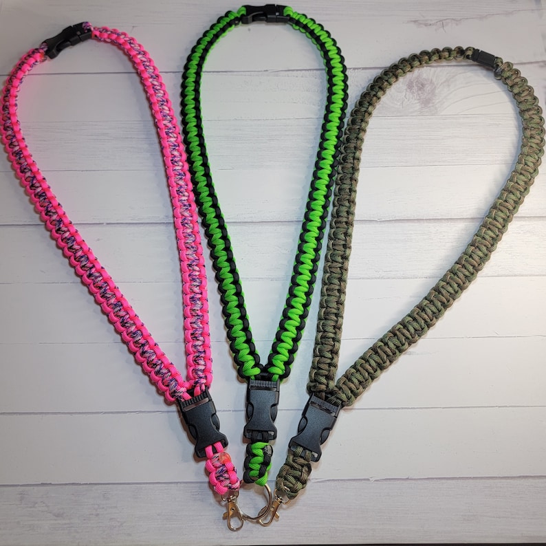 May include: Three paracord lanyards featuring a braided design with black plastic side-release buckles and metal swivel clips. The lanyards are colored in neon pink with purple accents, neon green with black, and a solid olive green camouflage pattern.