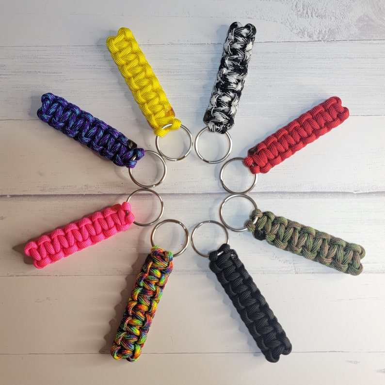 May include: Eight paracord keychains arranged in a circle, featuring various colours and patterns including solid yellow, black and white camo, solid red, green camo, solid black, rainbow, solid pink, and a blue and purple variegated cord design.