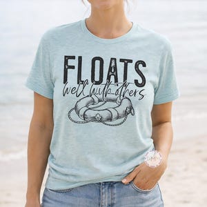 May include: Light blue t-shirt with the text "FLOATS well with others" and a graphic of a life preserver. The shirt is paired with denim shorts. The image is taken on a beach with a woman wearing sunglasses.