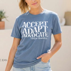 May include: A blue jean t-shirt with the words "ACCEPT, ADAPT, ADVOCATE" in white, along with "Autism Awareness" in a script font. The shirt is a solid color and has short sleeves.