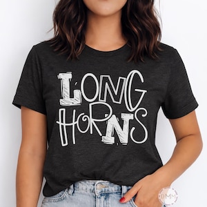 May include: Dark grey t-shirt featuring the white text "LONG HORNS" in a bold, outlined font. The short-sleeved shirt is made of a soft material. The shirt is worn by a person.