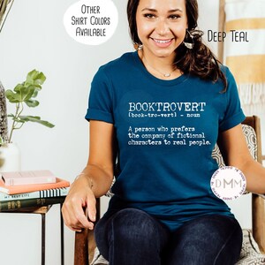 May include: Deep teal t-shirt with the word "BOOKTROVERT" and its definition printed in white. The shirt is a crew neck and has rolled-up sleeves. The text "OTHER SHIRT COLORS AVAILABLE" is in a white circle in the upper left corner.