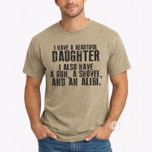 May include: A khaki short-sleeved crew neck t-shirt featuring black distressed text that reads: I HAVE A BEAUTIFUL DAUGHTER I ALSO HAVE A GUN, A SHOVEL, AND AN ALIBI. The shirt is paired with blue denim jeans, creating a casual look for everyday wear.