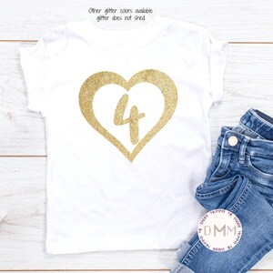 May include: White t-shirt with a gold glitter heart with the number 4 inside. The shirt is laying on a white wooden surface with a pair of blue jeans folded next to it.