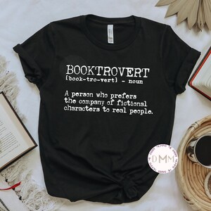 May include: Black t-shirt with the word "BOOKTROVERT" in white, along with a definition. The shirt is styled with open books, eyeglasses, and a coffee cup. The definition reads: "A person who prefers the company of fictional characters to real people."