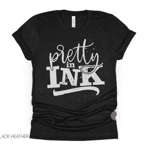 May include: A black heather short-sleeve t-shirt featuring the white text "pretty in INK" printed on the front. The word "pretty" is in a cursive script font, while "in INK" is in a bold, distressed block font with a horizontal line below.
