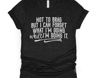 Op de afbeelding: Een zwart gemêleerd t-shirt met korte mouwen en witte tekst: NOT TO BRAG BUT I CAN FORGET WHAT I'M DOING WHILE I'M DOING IT. Het shirt heeft een ronde hals en een standaard pasvorm, met het kleurlabel BLACK HEATHER erboven weergegeven.