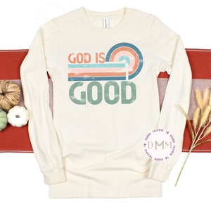 May include: Cream-colored long-sleeve shirt with the text "GOD IS GOOD" in a retro design. The design features a rainbow arch and horizontal stripes in shades of blue, orange, and pink. The shirt is displayed on a red and white striped surface.