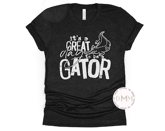May include: A black heather grey t-shirt with a white graphic that says "It's a great day to be a gator" with a cartoon alligator illustration.