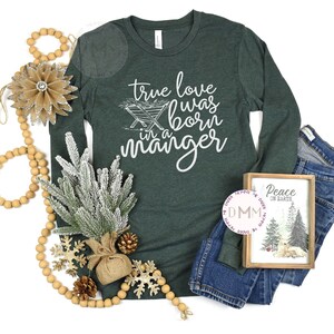 May include: A dark green long-sleeve shirt with the text "true love was born in a manger" in white. The image includes a framed picture with a winter scene, a strand of wooden beads, and decorative elements.
