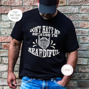 May include: Black t-shirt with the text "DON'T HATE ME BECAUSE I'M BEARDIFUL" in white. The design includes a stylized beard illustration. The shirt is paired with blue jeans and a black cap. Additional shirt colors are available.