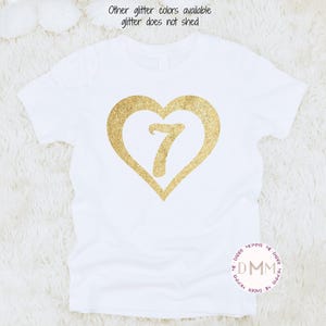 May include: White t-shirt with a gold glitter heart design and the number 7 inside. The text "Other glitter colors available glitter does not shed" is printed above the heart. A round logo with the text "DMM" is at the bottom.