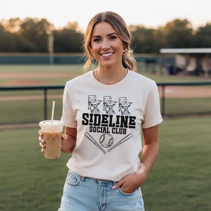 Op de afbeelding: Een cr&egrave;mekleurig T-shirt met korte mouwen en een zwarte grafische print van drie klapstoelen boven de tekst SIDELINE SOCIAL CLUB en een honkbal geflankeerd door twee gekruiste honkbalknuppels.