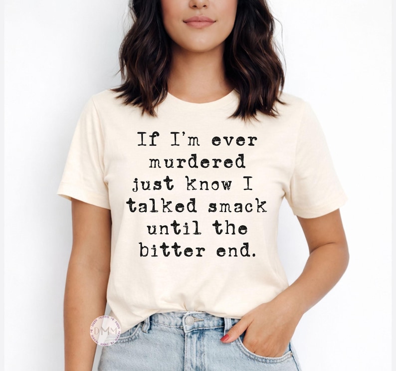 K&ouml;nnte beinhalten: Cremefarbenes T-Shirt mit schwarzem Text: "If I'm ever murdered just know I talked smack until the bitter end." Das kurz&auml;rmelige Shirt wird mit einer blauen Jeans getragen.