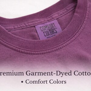 May include: A plum-colored t-shirt with a ribbed crew neckline and a purple interior neck label that reads COMFORT COLORS RING SPUN. Below the shirt, the text reads Premium Garment-Dyed Cotton and Comfort Colors in a dark serif font.