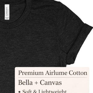 K&ouml;nnte beinhalten: Dunkelgraues T-Shirt mit hochgekrempelten &Auml;rmeln. Das Shirt hat einen Rundhalsausschnitt und ist aus einem weichen, leichten Material gefertigt. Das Etikett lautet "Premium Airlume Cotton", "Bella + Canvas" und "Soft & Lightweight".