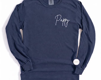 May include: A navy blue long-sleeve shirt featuring the word Pappy written in white cursive script on the upper right chest area. The garment has a crew neckline and a small circular white logo near the bottom right hem.