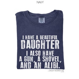 May include: A navy blue short-sleeved t-shirt with a distressed white graphic print that reads: I HAVE A BEAUTIFUL DAUGHTER I ALSO HAVE A GUN, A SHOVEL, AND AN ALIBI. The shirt features a Comfort Colors brand label at the inner neckline.