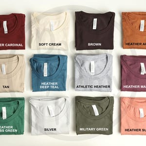 K&ouml;nnte beinhalten: Eine Sammlung gefalteter T-Shirts in verschiedenen Farben, darunter Heather Cardinal, Soft Cream, Braun und Heather Autumn. Jedes Shirt hat ein Etikett und ist mit seinem Farbnamen beschriftet. Die Shirts sind ordentlich auf einer wei&szlig;en Oberfl&auml;che angeordnet.