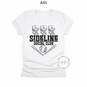 Op de afbeelding: Lichtgrijs t-shirt met een zwart grafisch ontwerp. Het ontwerp bevat drie klapstoelen, de tekst "SIDELINE SOCIAL CLUB", een honkbal en twee gekruiste honkbalknuppels. Een casual t-shirt voor sportfans.