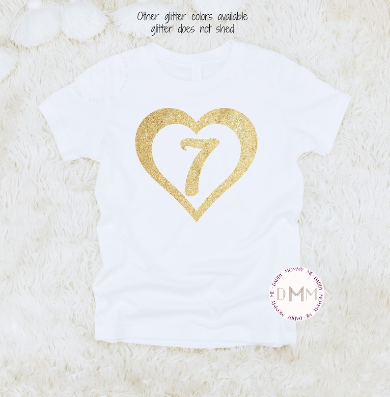 May include: White t-shirt with a gold glitter heart design and the number 7 inside. The text "Other glitter colors available glitter does not shed" is printed above the heart. A round logo with the text "DMM" is at the bottom.