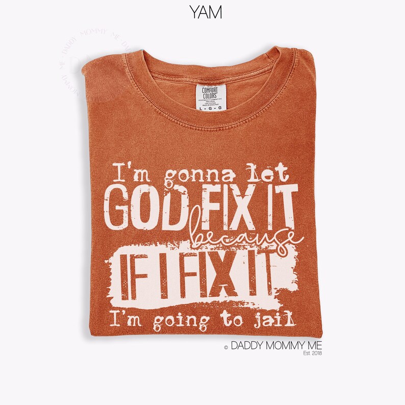 May include: A rust-colored short-sleeve t-shirt with a crew neckline. The front features white distressed-style text that reads: I'm gonna let GOD FIX IT because IF I FIX IT I'm going to jail. The shirt has a Comfort Colors brand label.