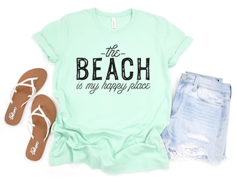 May include: A mint green short-sleeve t-shirt with the black text "the BEACH is my happy place" printed on the front, accompanied by a pair of brown flip-flops with white straps and light blue denim shorts with frayed hems and white lace trim.