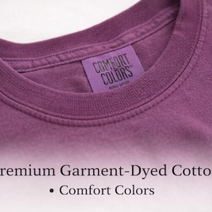 Puede incluir: Una camiseta color ciruela con cuello redondo acanalado y una etiqueta interior morada que dice COMFORT COLORS RING SPUN. Debajo de la camiseta, el texto dice Premium Garment-Dyed Cotton y Comfort Colors en una fuente serif oscura.