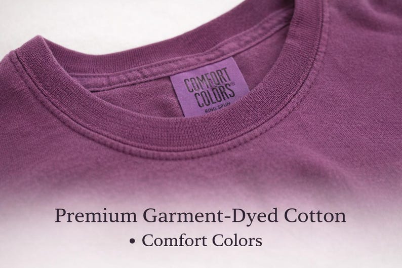 Puede incluir: Una camiseta color ciruela con cuello redondo acanalado y una etiqueta interior morada que dice COMFORT COLORS RING SPUN. Debajo de la camiseta, el texto dice Premium Garment-Dyed Cotton y Comfort Colors en una fuente serif oscura.