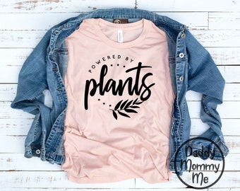 May include: A pale pink t-shirt with the text "POWERED BY plants" in black script and a leaf design. A denim jacket is draped over the shirt, set on a white wooden surface.