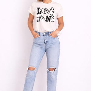 May include: A cream-coloured t-shirt with the text "LONG HORNS" in black, worn with light blue ripped jeans. The model is wearing white trainers. The overall style is casual and comfortable.