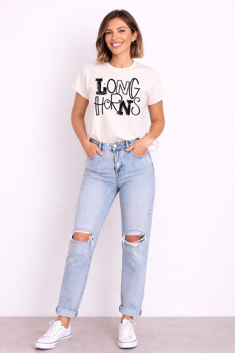 May include: A cream-coloured t-shirt with the text "LONG HORNS" in black, worn with light blue ripped jeans. The model is wearing white trainers. The overall style is casual and comfortable.