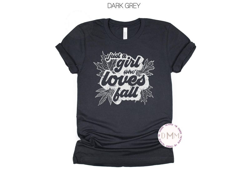 May include: Dark grey t-shirt with the text "just a girl who loves fall" in white, surrounded by leaf illustrations. The shirt is a solid color and has a crew neck.