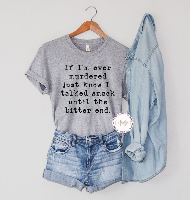 K&ouml;nnte beinhalten: Ein graues T-Shirt mit dem Text "If I'm ever murdered just know I talked smack until the bitter end." Das Shirt wird mit Jeansshorts und einem hellblauen Jeanshemd kombiniert. Das T-Shirt h&auml;ngt an einem Holzb&uuml;gel.