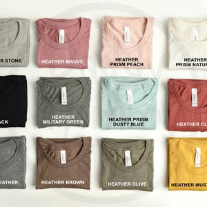 K&ouml;nnte beinhalten: Eine Auswahl an gefalteten T-Shirts in verschiedenen Farben, darunter Heather Stone, Mauve, Pfirsich und Natur. Jedes Shirt hat ein Etikett mit dem Farbnamen. Weitere Farben sind Schwarz, Milit&auml;rgr&uuml;n, Dusty Blue und Clay.