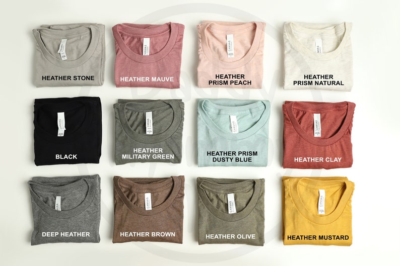 K&ouml;nnte beinhalten: Eine Auswahl an gefalteten T-Shirts in verschiedenen Farben, darunter Heather Stone, Mauve, Pfirsich und Natur. Jedes Shirt hat ein Etikett mit dem Farbnamen. Weitere Farben sind Schwarz, Milit&auml;rgr&uuml;n, Dusty Blue und Clay.