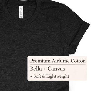 K&ouml;nnte beinhalten: Dunkelgraues T-Shirt mit hochgekrempelten &Auml;rmeln. Das Shirt hat einen Rundhalsausschnitt und besteht aus einem weichen, leichten Material. Das Etikett lautet "Premium Airlume Cotton" und "Bella + Canvas".